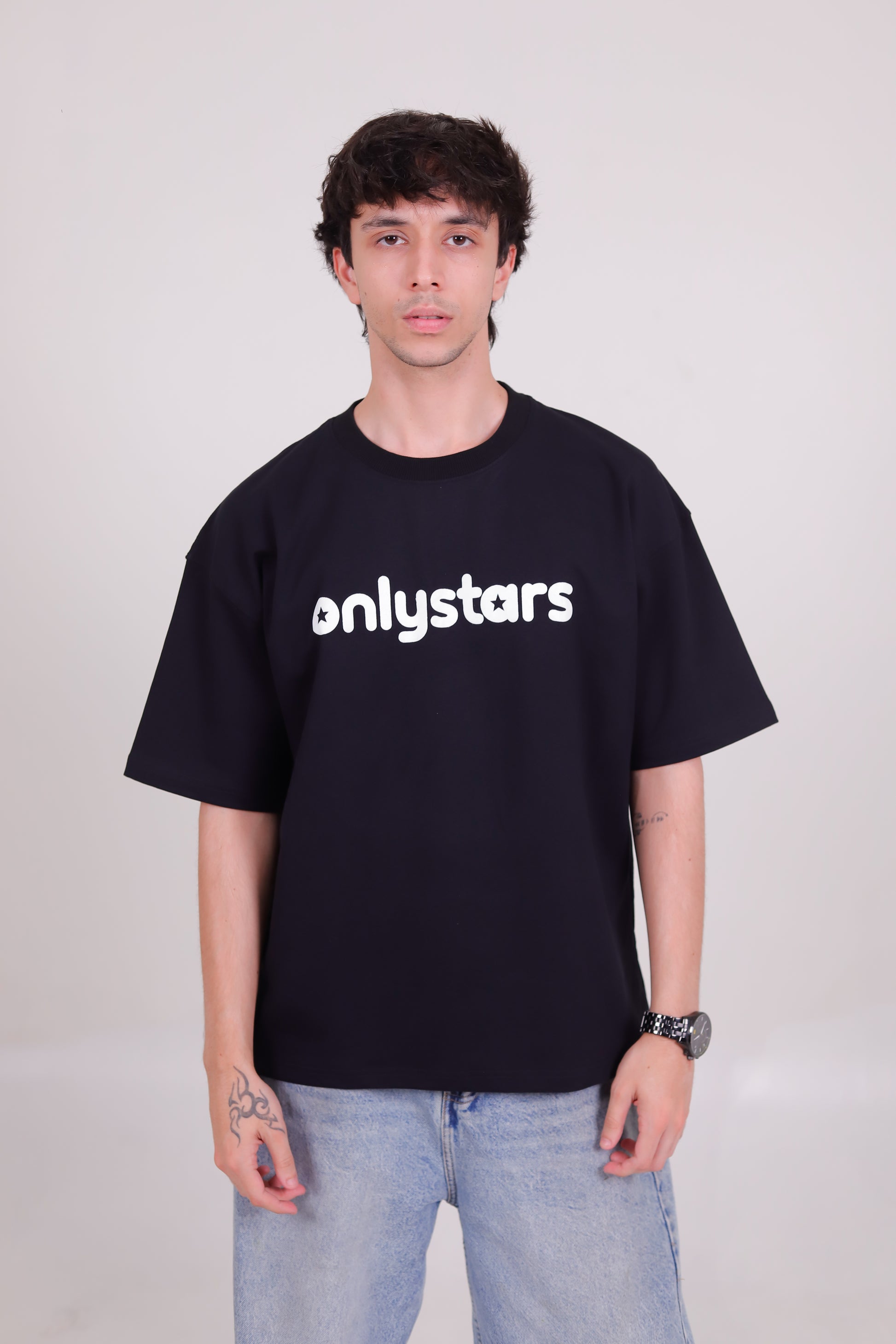 A man wearing a black crewneck t-shirt with the word 'onlystars' printed in white across the chest, paired with light blue jeans, standing against a light gray studio background.